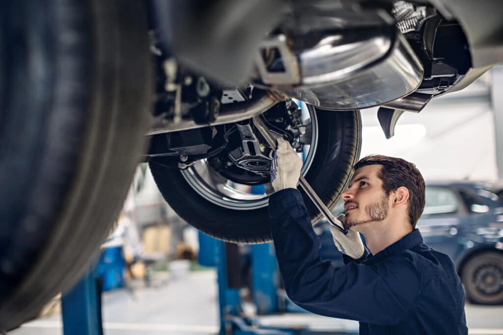 Mécanicien auto en action : Réparation sous la voiture en atelier Mécanicien en gants travaillant sous une voiture soulevée dans un atelier de réparation, utilisant un outil sur le dessous du véhicule.