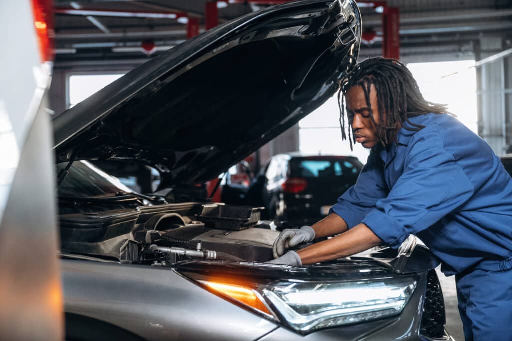 Mécanicien afro-américain concentré, en uniforme bleu et gants, travaillant sur le moteur ouvert d'une voiture dans un garage.
